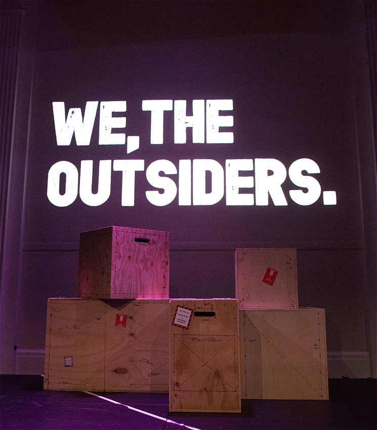 A collection of wooden boxes piled next to and on top of each other. Behind them text is projected against a back wall reading: 