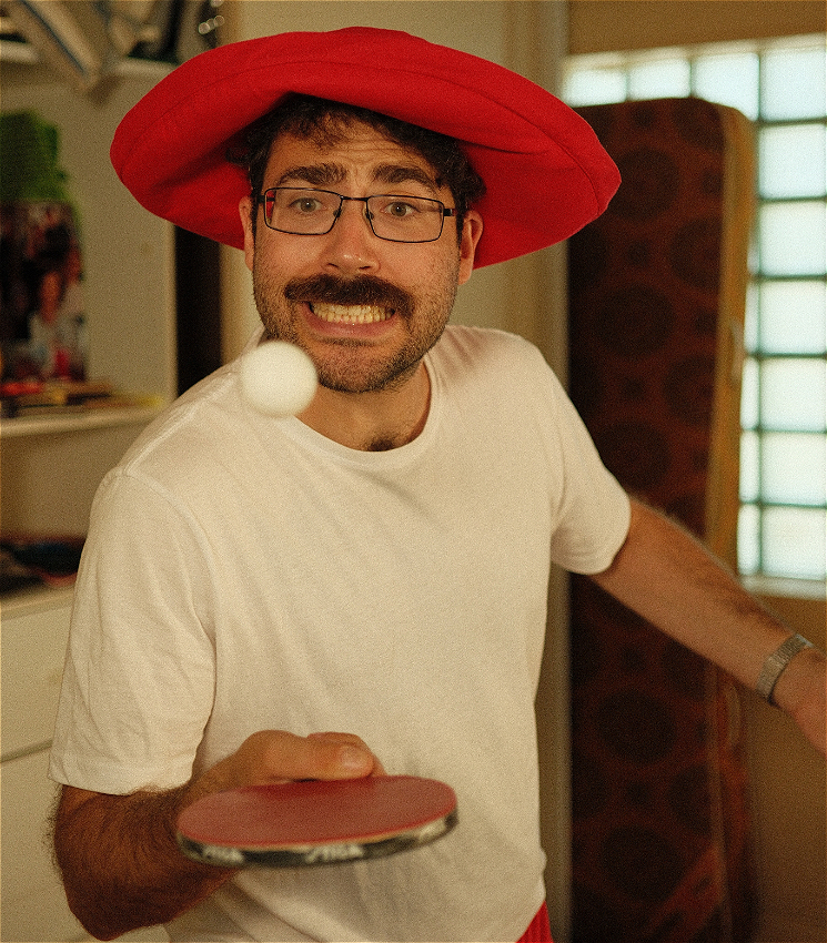 A man in his 20s wearing a big red dumb hat is bouncing a table tennis ball on a bat. He has a grimace plastered across his face.