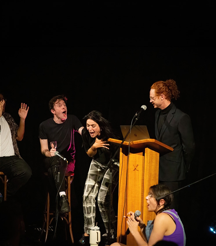 The image depicts 5 individuals on stage, all 5 of them are seated on stools with multiple microphones pointed at them. The man on the left seems to be leaning back in shock, the man beside him also appears shocked, touching the shoulder of the woman next to him. The woman beside him is slightly crouching clutching her chest in shock. The two hosts are on the right, one is standing behind a lectern on the stage, and a woman is seating on the stage beside him also holding a microphone.