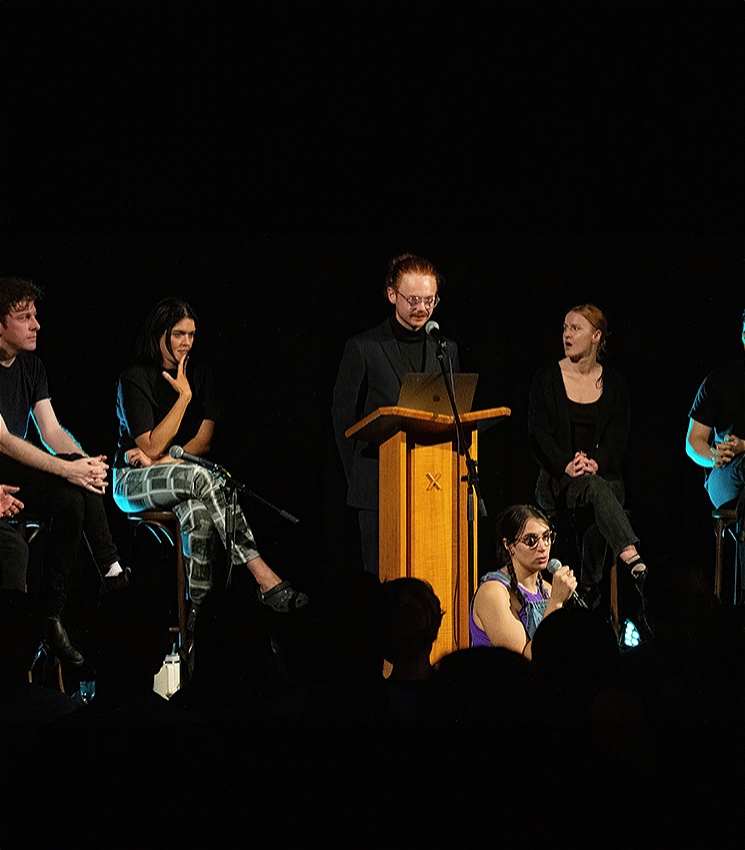 The image shows 5 people on a darkly lit stage. The central host is man dressed in all black standing behind a lectern with a microphone pointed towards him. There is a woman who is also hosting seated beside him on the floor of the stage holding a microphone. The 3 other people are seated on stools all listening intently.