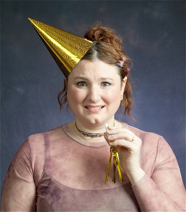 A medium close up shot in the style of a 90s school picture - dark grey gradiated background and that trademark center glow. In the foreground is an adult woman dressed like a teenager from 1999 - wearing a long sleeved mesh top with a black singlet underneath, a choker necklace, and butterfly clips in her auburn hair, which is in a 90's era up-do: straight at the front - curly at the back. She has a gold party hat askew on her head and holds a gold party blower, and while she's technically smiling, underneath she looks terrified.