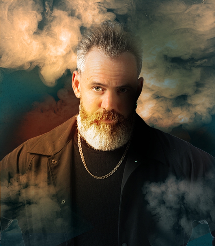 A moody portrait of a man with grey hair and beard. He is wearing a black shirt jacket over a black t-shirt with a gold chain necklace. He is in front of a dark background filled with backlit smoke that's also filtering in front of him.