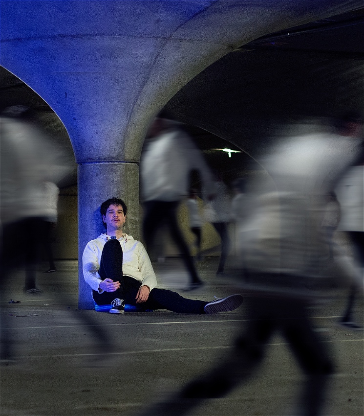 A person in a white sweater with black jeans and converses sits leaning against a large concrete pillar. They are staring at the camera with a knowing look. The pillar is lit in a blue light and arches out into the ceiling. Around the pillar are blurs of the same person in motion.