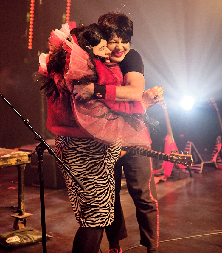 On a stage, two people hug. The left is a woman with black hair and vibrant red-and-blue makeup. On the right is a man with black hair and bright red lipstick, who is wearing a band t-shirt.