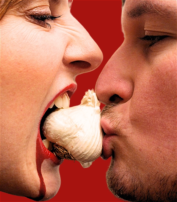 A man and women's facial side profile, both kissing a bulb of garlic. The woman's face has blood dripping from her lip. Their faces are on front of a red background.