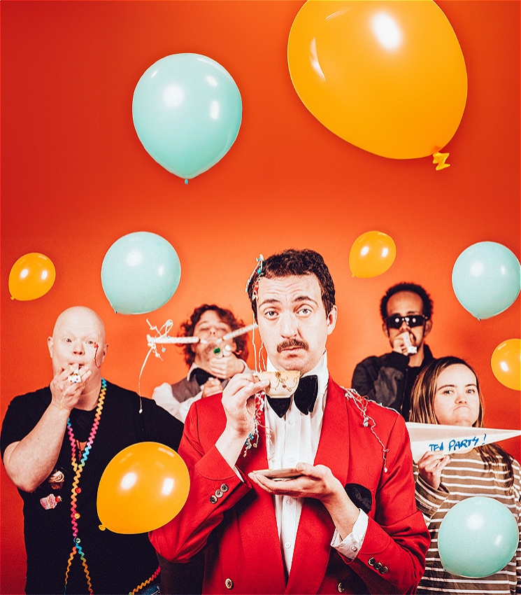 A man in a red blazer and black bow tie delicately holds a tea cup and saucer in front of an orange background as an outrageous party is in full swing behind him. The four people behind him are holding streamers and party poppers, and the scene is filled with orange and blue balloons.