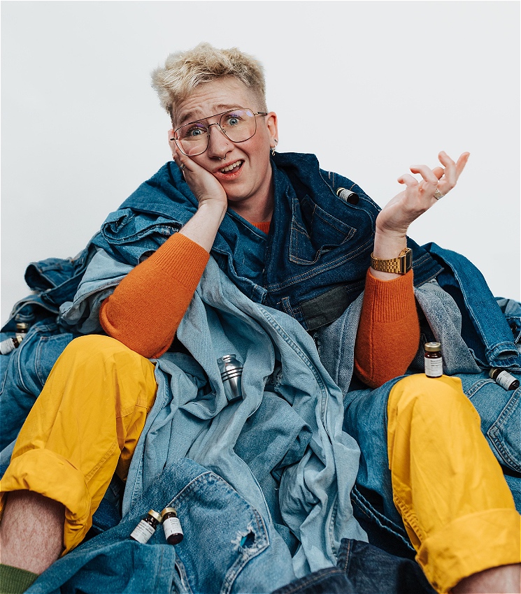 Eddie sits upright on the floor partially consumed by a big pile of blue denim jeans, with their legs, arms and head emerging out. Scattered amongst the jeans are seven empty vials of Testosterone and one small silver keepsake urn. 
Eddie is white, they have a cropped bleached blonde mullet, big wire frame glasses, and multiple piercings in their nose. 
They have a confused, overwhelmed and slightly amused expression on their face.