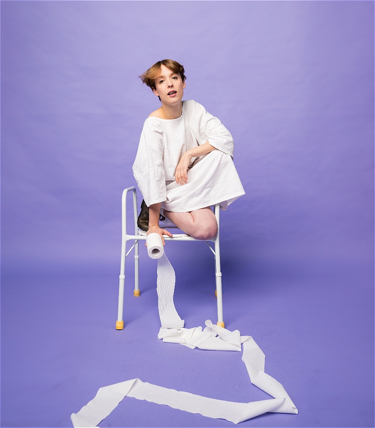 Hayley Edwards crouches on an accessible toilet seat wearing a white hospital gown and holding a toilet roll that's spreading over the floor. The background is a purple-lavender colour.