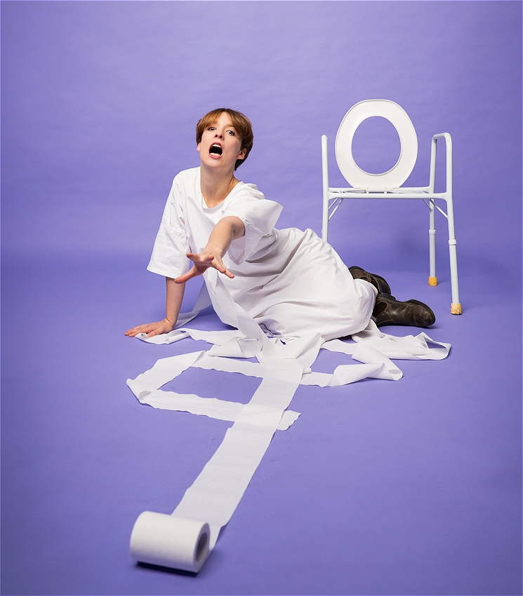 Hayley Edwards reaches out for a toilet roll on the floor with a desperate expression. They wear a white hospital gown, the background is a purple-lavender colour and there's an accessible toilet seat in the background.