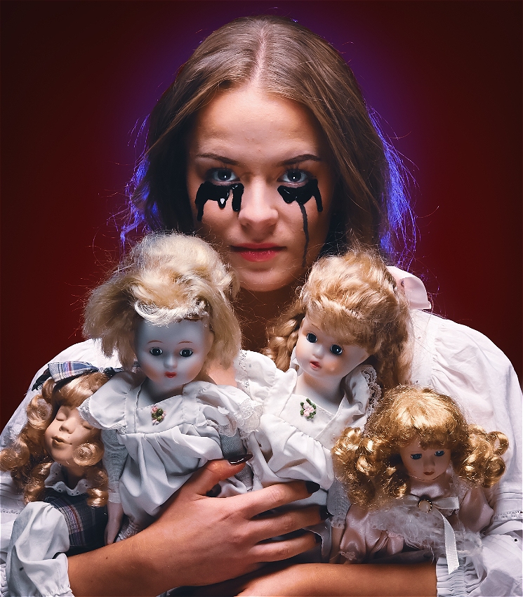 A young woman in white holds four vintage dolls as black liquid falls from her eyes. The dolls are clutched to her chest and she looks at the camera with an unflinching expression.