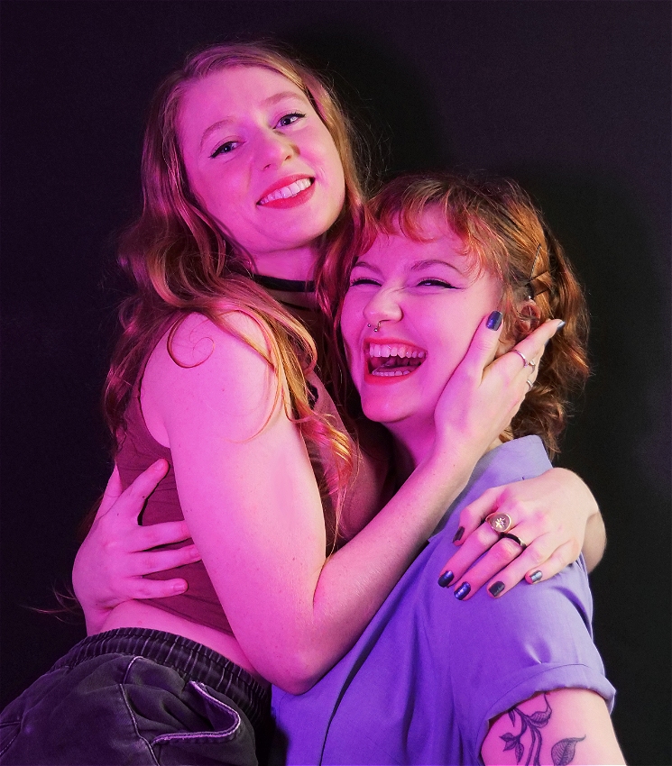 Two people pressed closely together and smiling happily illuminated by pink lighting, creating a warm and loving atmosphere, in front of a black background. The person on the left has long, wavy hair and is wearing a singlet. They are being held up by the person on the right, who has short, curly hair and is wearing a collared shirt. The person on the right has their arms around the person on the left's waist, lifting them slightly off the ground, while the person on the left has their hand gently placed on the other’s cheek.