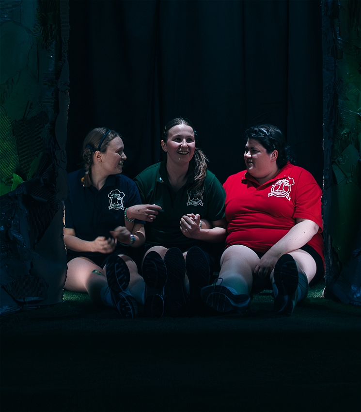 Three people sit tightly enclosed in a hollow tree made out of cardboard. They wear school polo shirts coloured blue, green and red with a school logo on them. They are holding hands and have excited looks on their faces.