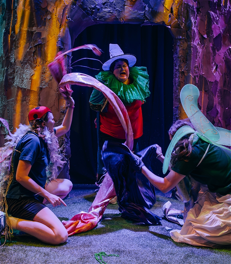 Three performers wear colourful clothes in front of a large tree made out of cardboard. One performer is standing in the middle wearing a witch's hat and cloak over a red polo shirt in mid-scream. The other two performers kneel on the ground wearing a mix of fancy dress pieces over their dark school polo shirts. They are giggling and waving coloured fabric around the standing performer - pretending to 'burn the witch'.