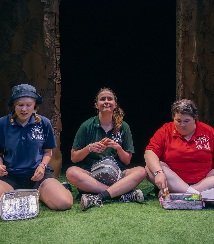 Three performers sit on astroturf in front of a cardboard tree, smiling. They wear polo shirts of different colours with a primary school logo on each. One performer wears a bucket hat, the other two wear headbands. They have colourful school lunchbags and are eating recess snacks.