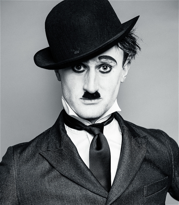 A medium black and white close up of a young caucasian man dressed as Charlie Chaplin. He is wearing a tight black suit jacket with a white shirt and a black tie and he is wearing a tilted bowler hat on his head, almost covering his right eye. He has Chaplin style makeup with raised black eyebrows, heavy black eyeliner and a small moustache. He looks straight into the camera.