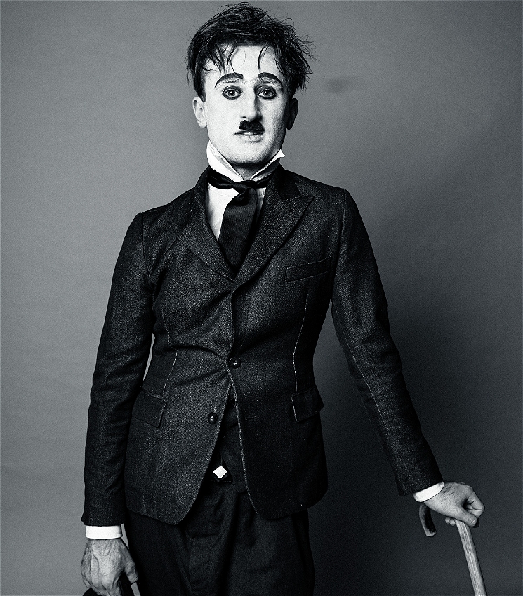 A medium black and white shot of a young caucasian man dressed as Charlie Chaplin. He is wearing a tight black suit jacket, a black vest, a white shirt with a black tie and baggy black pants. He has Chaplin style makeup with raised black eyebrows, heavy black eyeliner and a small moustache. He leans on his left hand in which he holds a wooden cane and in his right hand, he holds a bowler hat. He looks straight into the camera with a casual smile.