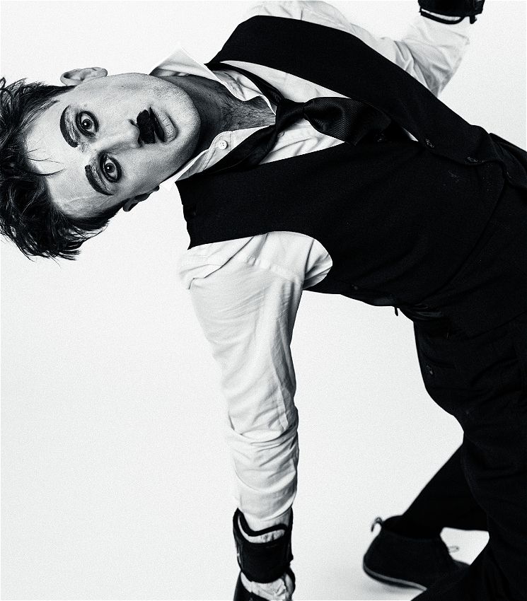 A medium black and white close up of a young caucasian man dressed as Charlie Chaplin. TheHe is dressed in a white shirt, a black vest and a black tie with baggy black trousers and black shoes and he is wearing boxing gloves. The shirt has a few buttons undone and the tie is lose. He has Chaplin style makeup with raised black eyebrows, heavy black eyeliner and a small moustache. He is in a back bend, as if just punched in the face and he looks straight into the camera with a slightly stunned expression on his face.