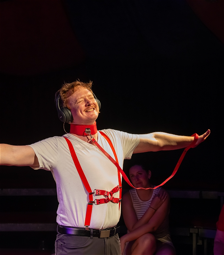 A man wearing a red leather harness over a white tshirt with black headphones on has arms outstretched and is smiling with joy as if listening to something lovely.