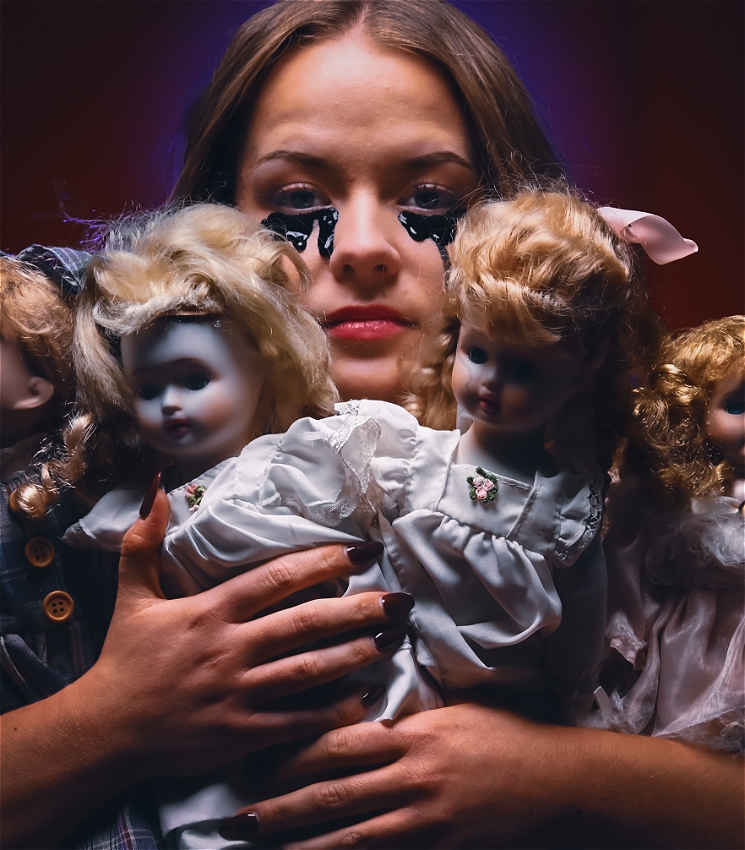 Close up on a young woman in white as she four vintage dolls over her face so as to partially obscure it. Black liquid leaks from her eyes as she holds the dolls and looks at the camera through their heads