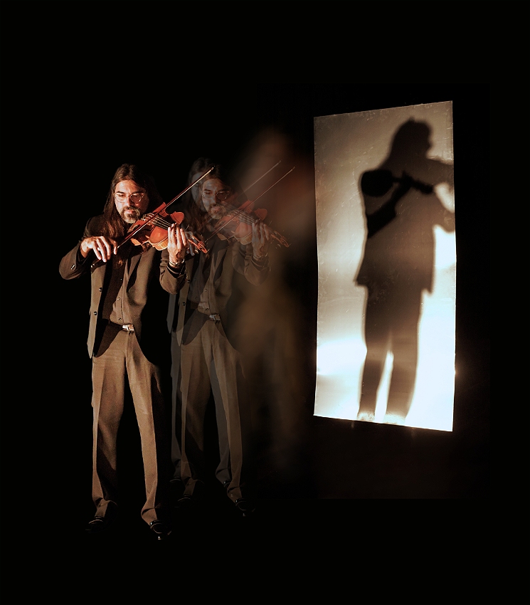 A person playing a viola in a dark room, with a large metal sheet reflecting both their image and a projection of their shadow.