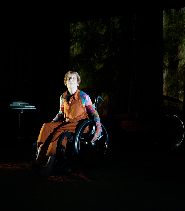 A photo of Melinda on stage, sitting in her wheelchair. She has short, sandy blonde hair, pale skin and is looking up towards the ceiling. She is wearing an orange jumpsuit and a long-sleeved top underneath with an abstract pattern in blue, pink, purple and orange. She sits in front of a dark, projected background image of a forest with tall trees. There is a typewriter on a stand to the left and a partially visible drumkit to the right.