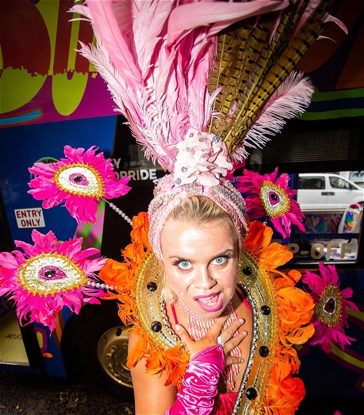 A blonde woman wearing a very colourful, feathery and sparkly burlesque/cabaret outfit and head dress leans toward the camera, hand on her chest