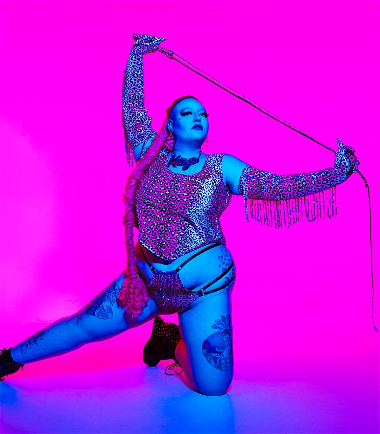 The image shows a person posing for a photo in a dynamic and powerful way. They are wearing a leopard print outfit and holding a whip. The background is a vibrant pink and blue, adding to the dramatic and colourful feel of the image.