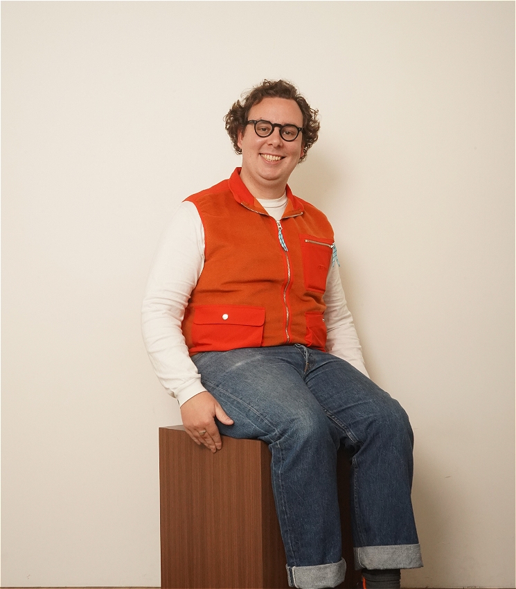 Darcy is sitting on a wooden box at a 45 degree angle to the camera. He has a precocious smile, curly hair and dark thick-rimmed glasses. He is wearing a bright orange vest over a white long-sleeve t-shirt and light blue jeans.