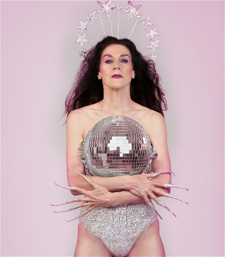 An image of a woman holding a mirror ball in her arms, as she stands against a silver-pink background. She is wearing a shimmering silver corset, has long, silver taloned fingernails, long dark hair, curled eyelashes, sparkling pink eye shadow and a closed mouth wearing dark pink lipstick. Silvers stars on slender silver sticks appear to be forming a crown above her ahead.