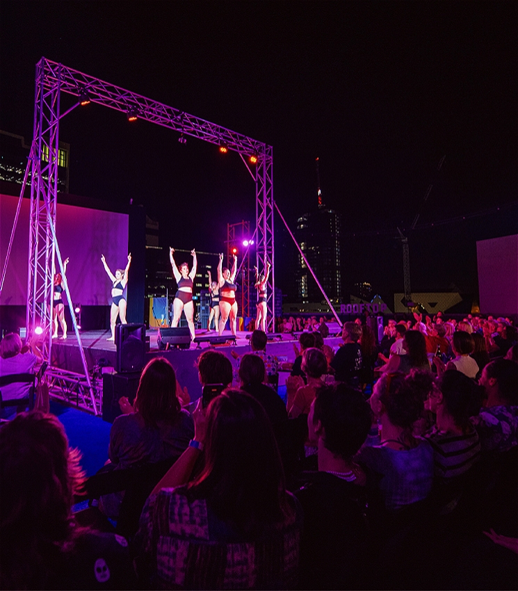 The cast of YUCK Circus receive applause from their sold out audience, at the end of their show. The cast are onstage, with their arms in the air, the couple of hundred-strong audience seated around them cheering. The stage is outside on a rooftop, under a cityscape.