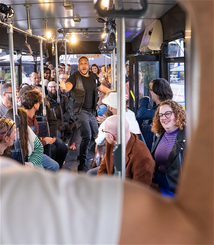A man in a SWAT style uniform stands onboard a bus surrounded by passengers. He looks tense.