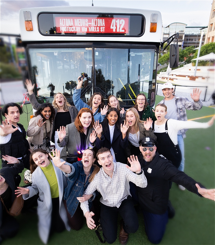 A group of audience members stand in front of a red and white bus. They have just seen a performance of 
