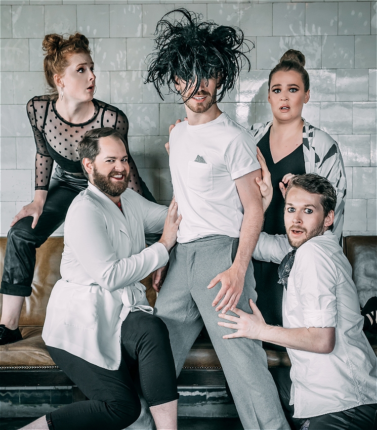 Four performers wearing black and white surround another performer with a wig on with black messy hair covering his face.