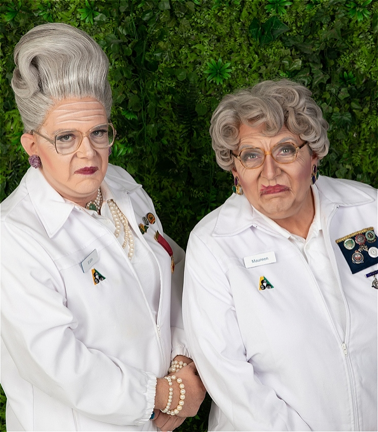 Two drag queens dressed as very old women are wearing white lawn bowls uniforms standing in front of a shrub smirking at the camera.
