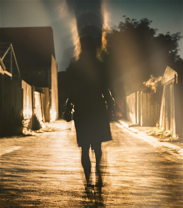 A blurred photograph of a person walking down a suburban street at nighttime with their back to the viewer.