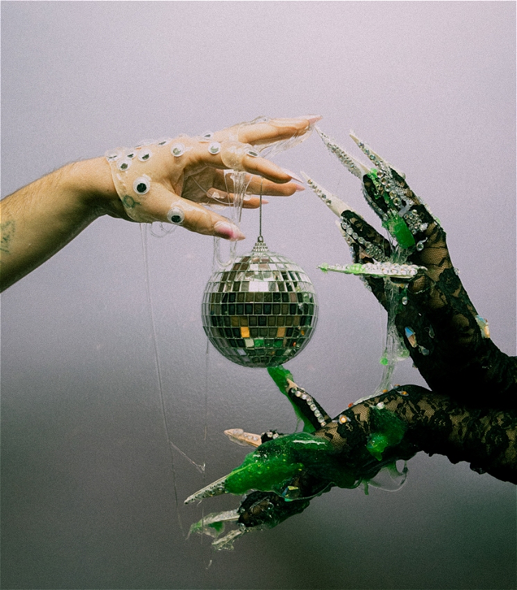 Three hands decorated with long nails, googly eyes and diamonds extend out to one another covered in green and clear slime.