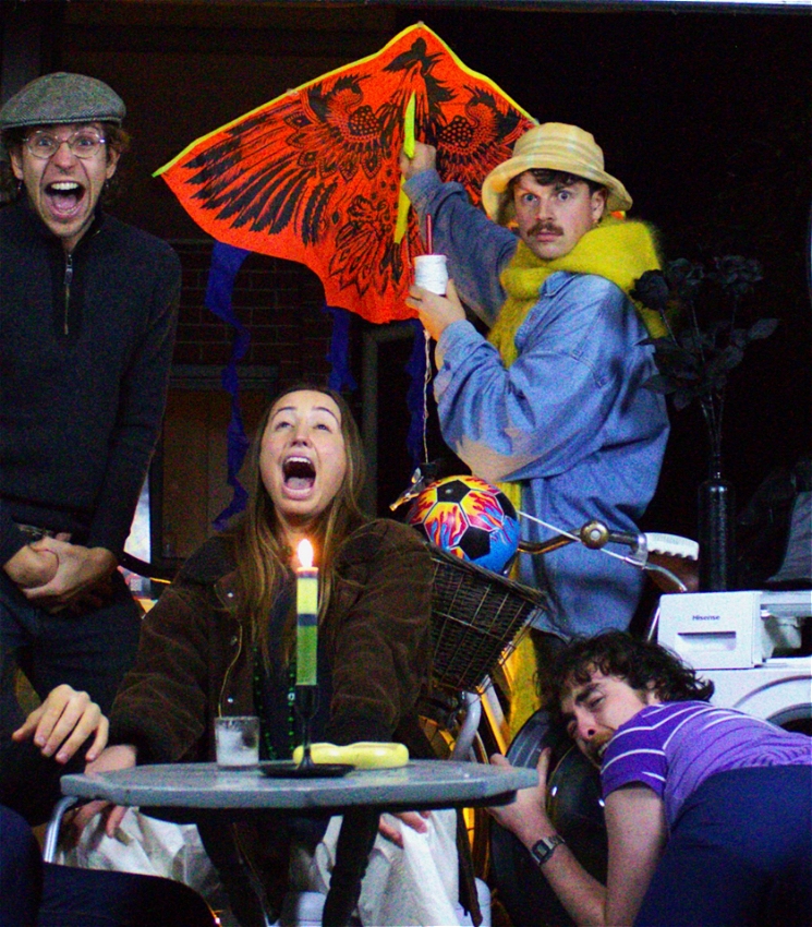 The image features chaotic snapshot of 4 people in a cluttered garage at night time, with several props featuring. A man dressed in an oversized blue-denim jacket, thick yellow scarf and a yellow hat stares intensely into the camera. He is holding a bright orange kite with black print of a bird with it's wings spread. Around him are three other people shouting at each other and at the camera with their mouths wide open. There's table with a lit green candle, a washing machine and an inflated ball with blue, black and yellow panel around these characters.