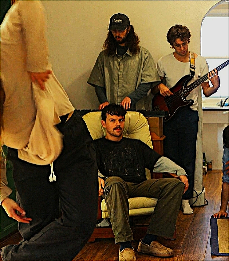 In the middle of the picture a young white man with short brown hair and a moustache sits in a living room on a white chair staring at a young woman dancing who is in the very foreground of the image. Behind the young man are two musicians, a tall keyboard player with long hair and a bass player. You can also just see the arm of another dancer entering from the doorway at the back.