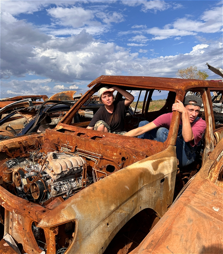 Two boys lean out of a rusted and derelict car, looking to camera.