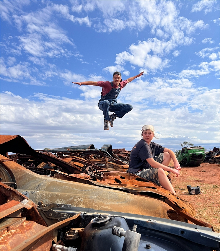 Two boys in a junk yard, filled with burnt out cars. One looks at camera in the foreground whilst the other jumps in the air behind.