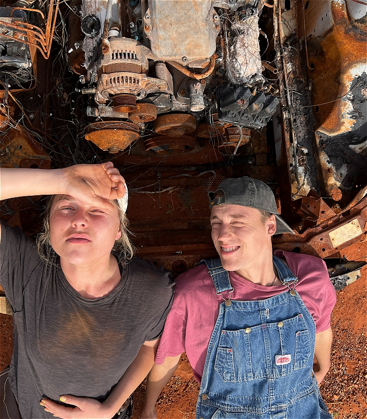 Two boys squint up at camera, from the bonnet of a rusted and burnt out car. The boy on the left, wearing a dark shirt and cap, looks up shielding his eyes from the sun. The boy on the right, in a red shirt and overalls, squints his eyes.