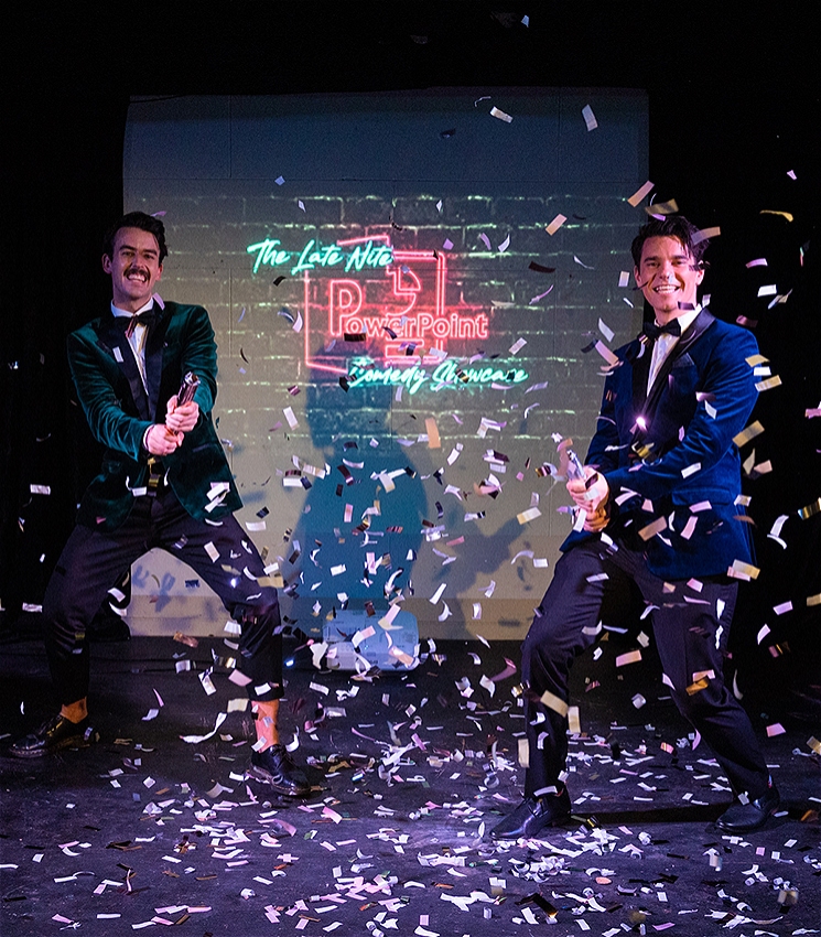 Two men in velvet suits are holding confetti cannons, they're standing front of a projector screen smiling while confetti rains down in front of them