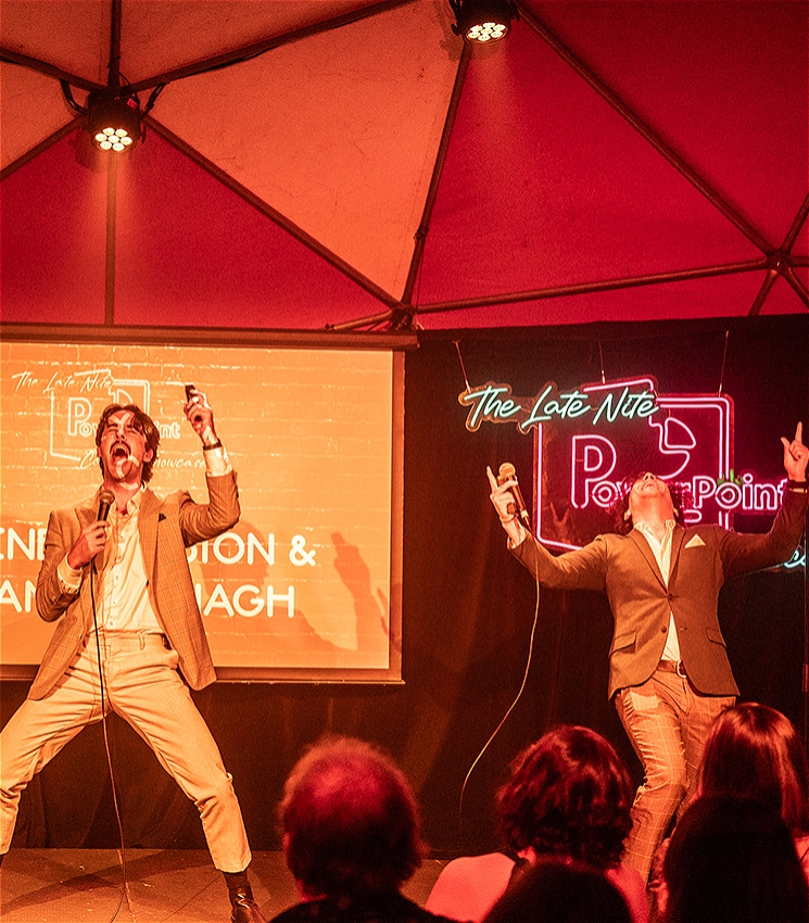 Two men wearing suits stand on stage yelling with their hands flung in the air. They hold micophones and presentation clickers. In the background there is a projector screen and a neon sign hanging at the back of the stage.