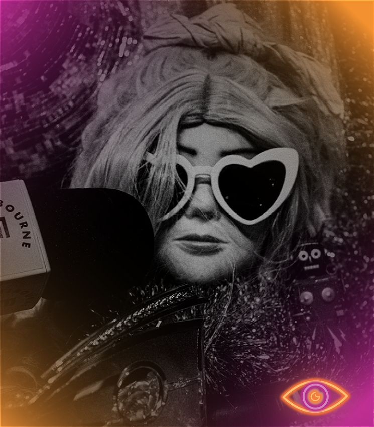 A mannequin head wearing a blonde wig and heart shaped sunglasses sits amongst bits of fabric and a disco ball. A microphone presses towards its mouth. The microphone has the words 