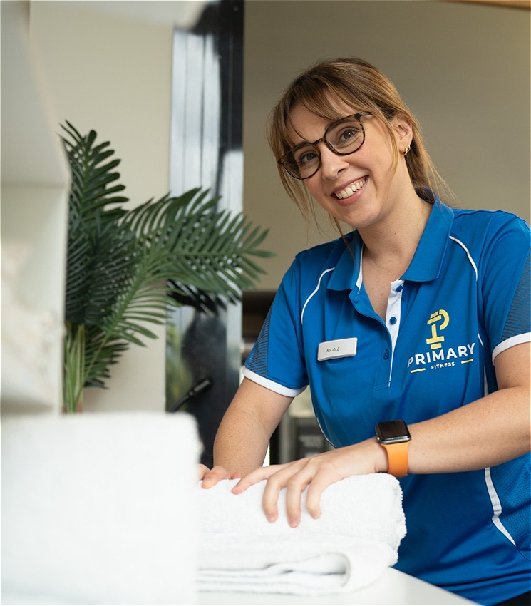 The image shows a woman with light brown hair and glasses, smiling. She is wearing a blue polo shirt with a 'Primary Fitness' logo and is folding white towels. In the background, there is a plant and a window, suggesting an indoor setting. She also wears an orange-strap smartwatch on her wrist.