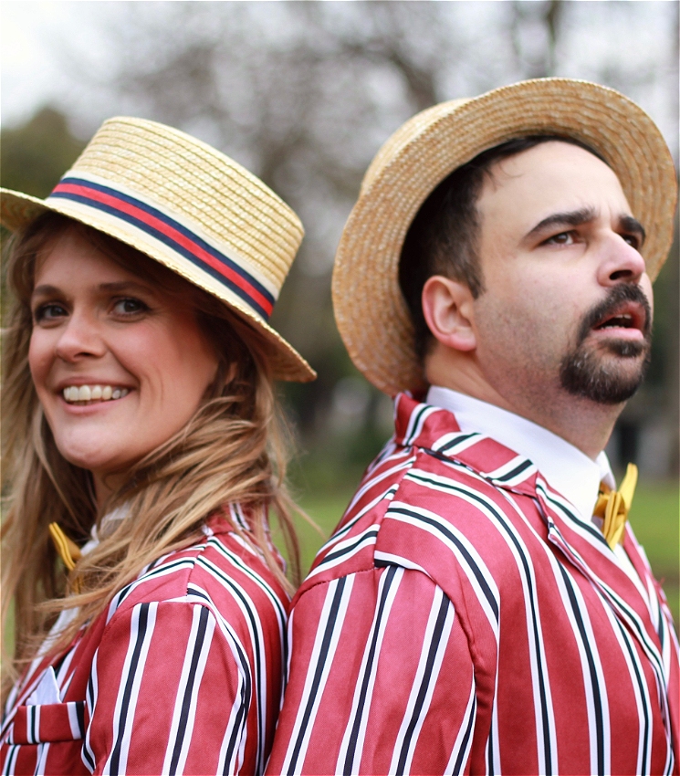 In a park, a man with facial hair and a woman with long blonde hair stand back-to-back. They are wearing matching striped red suits with yellow bow ties and flat straw hats. She has a cheeky smile and he looks somewhat frustrated.