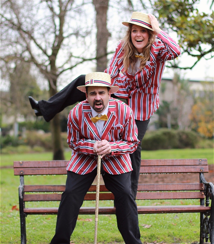 In a park, a man with facial hair hunches over a walking stick in pain, while a woman with long blonde hair gleefully rests atop his back, with one foot in the air. They are wearing matching striped red suits with yellow bowties and flat straw hats.