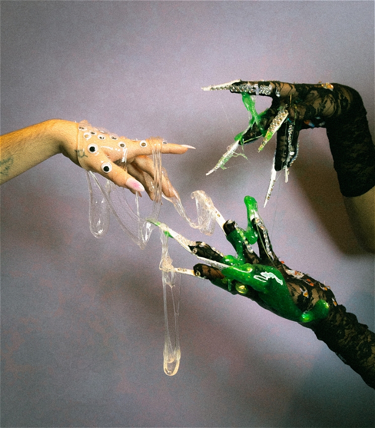 One hand decorated with long nails, googly eyes and diamonds extend out to another two hands in black gloves, covered with green slime. Clear slime is hanging from all three hands.