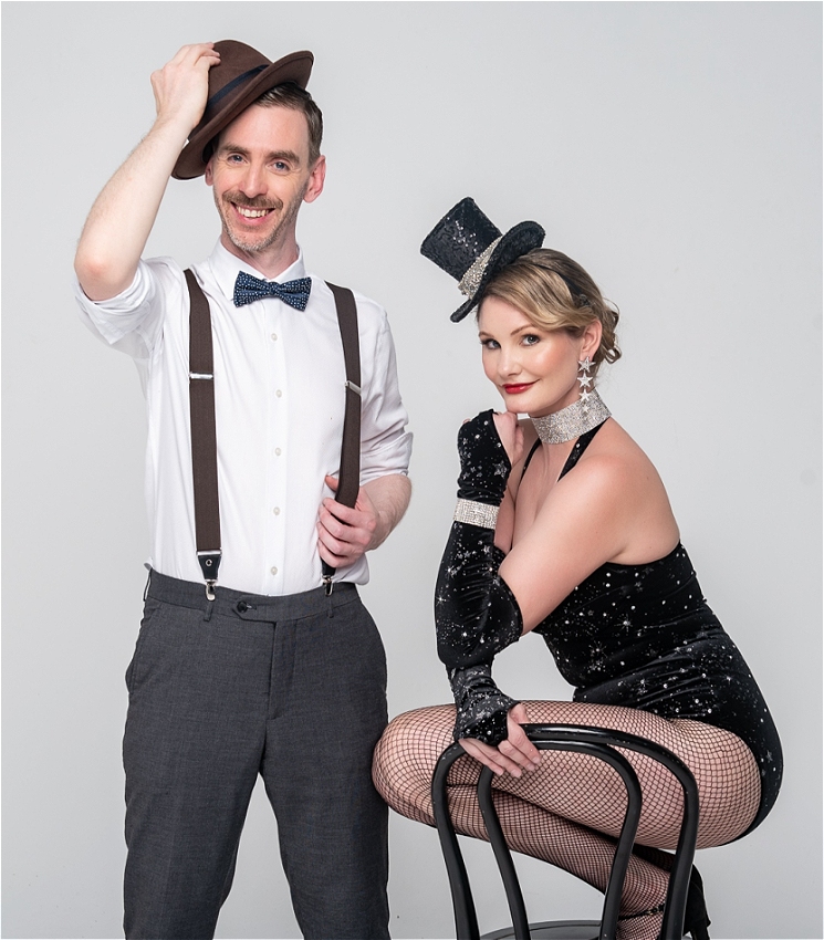 Greg, a white man in his 40s is standing holding a hat with a white shirt and bow tie, and Isabella, a woman in a black sparkly leotard and hat with a diamante choker, is standing crouched on a black bentwood chair next to him.