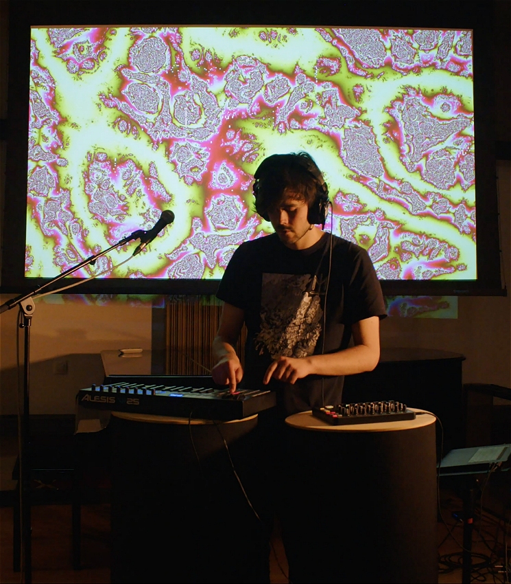 A man is standing in front of a microphone and electronic music equipment. He is wearing headphones and a dark shirt. Behind the man is a visual projection of green and pink abstract patterns.